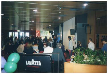 COL photo of a large group of people standing around several large tables. One woman is standing in the centre of the frame,  near a large freestanding blue and gold banner, some balloons, and a speaker on a tall stand. On top of the tables, there are cups and sauces and other miscellaneous items. In the background, there are several large windows along one wall. In the foreground, there are some balloons to the left of the frame, a Lavazzo branded barrier/room divider in the centre of the frame, and a large circular planter containing several potted plants to the right of the frame.