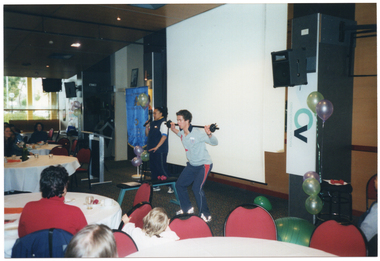 COL photo of a woman speaking into a handheld cordless microphone while a man demonstrates a weightlifting exercise. He is holding a small set of barbells across the back of his shoulders while bending his knees slightly. Behind them, there is a large projector screen, a lectern, a vertical blue and gold banner, and some balloons. In the foreground, groups of people are sitting at several large round tables.