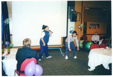 COL photo of a woman speaking into a handheld cordless microphone while a man demonstrates a weightlifting exercise. He is bending forward while holding a small set of barbells across in front of his slightly bent knees. Behind them, there is a large projector screen, a speaker on a tall stand, and a few balloons. In the foreground, a few people are sitting at large round tables.