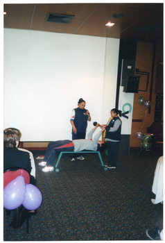 COL photo of two woman standing around a man who is lying on his back on an exercise bench. One of the women is speaking into a handheld cordless microphone while the man demonstrates a weightlifting exercise. He is holding a small set of barbells high above his head, assisted by the other woman. Behind them, there is a large projector screen, a speaker on a tall stand, and a few balloons. In the foreground, a few people are sitting at large round tables.