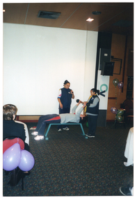 COL photo of two woman standing around a man who is lying on his back on an exercise bench. One of the women is speaking into a handheld cordless microphone while the man demonstrates a weightlifting exercise. He is holding a small set of barbells high above his head, assisted by the other woman. Behind them, there is a large projector screen, a speaker on a tall stand, and a few balloons. In the foreground, a few people are sitting at large round tables.