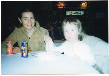 COL photo of a woman and young girl. They are sitting at a table, and the woman is smiling. In the background, a large sign advertising catering packages depicts the monsu.org website address in the bottom right corner.