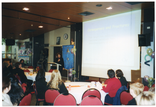 COL photo of a woman, standing at a lectern, speaking into a microphone. There is a large projector screen to her left, on which a slide titled 'Osteoporosis and Fractures' is being projected. Behind her is a freestanding blue and gold Arthritis Victoria/Osteoporosis Victoria banner, some balloons, and a speaker on a tall stand. To the right of the frame, there is an Osteoporosis Victoria banner and some balloons. In the background, there is a large display board on the wall, covered in posters. In the foreground, groups of people are seated at several large round tables.