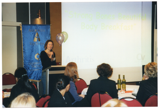 COL photo of a woman, standing at a lectern, speaking into a cordless microphone.  There is a large projector screen to her left, with a slide displaying the text, "Strong Bones Beautiful Body Breakfast". Behind her, there is a freestanding blue and gold Arthritis Victoria/Osteoporosis Victoria banner. In the foreground, there are several people sitting at large round tables.