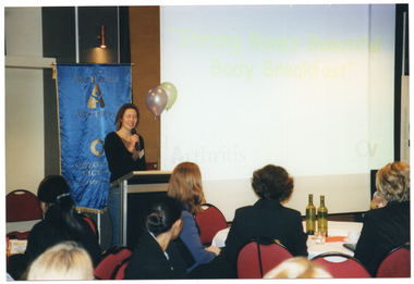 COL photo of a woman, standing at a lectern, speaking into a cordless microphone.  There is a large projector screen to her left, with a slide displaying the text, "Strong Bones Beautiful Body Breakfast". Behind her, there is a freestanding blue and gold Arthritis Victoria/Osteoporosis Victoria banner. In the foreground, there are several people sitting at large round tables.