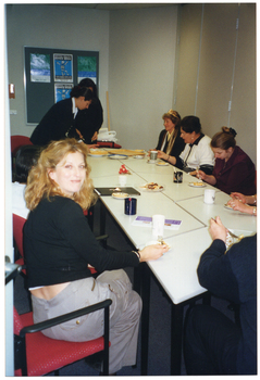 COL photo of a small group of people sitting around some rectangular tables. There are cakes and mugs on the table. One woman is wearing a plastic crown on her head.