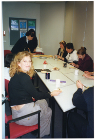 COL photo of a small group of people sitting around some rectangular tables. There are cakes and mugs on the table. One woman is wearing a plastic crown on her head.