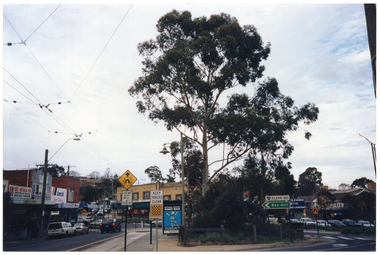 COL photo of a large advertisement on the side of a bus shelter. In the foreground, there are several road traffic signs. Around and behind the bus shelter, there are various retail stores.