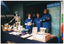 COL photo of three women, wearing bright blue t-shirts displaying the "Arthritis Victoria" text and logo. They are standing behind a long rectangular table covered with a dark blue table cloth. One woman is holding a round ball printed with 'Mitre Tournament' text and logo. Also behind the table, alongside the women, there is a large plastic skeleton on a stand wearing a blue cap and scarf. On the table in front of them, there are various examples of fresh and packaged food items, all labelled, and a range of brochures/fact sheets. Behind them, there is a freestanding blue and green Arthritis Victoria banner.