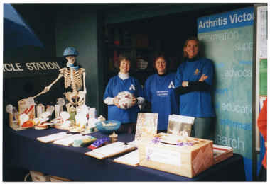 COL photo of three women, wearing bright blue t-shirts displaying the "Arthritis Victoria" text and logo. They are standing behind a long rectangular table covered with a dark blue table cloth. One woman is holding a round ball printed with 'Mitre Tournament' text and logo. Also behind the table, alongside the women, there is a large plastic skeleton on a stand wearing a blue cap and scarf. On the table in front of them, there are various examples of fresh and packaged food items, all labelled, and a range of brochures/fact sheets. Behind them, there is a freestanding blue and green Arthritis Victoria banner.