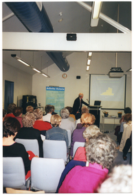 COL photo of a man standing behind a lectern. He is turning around to look at a large slide projector screen behind him. On the screen, there is a slide depicting a graph. To the left of the frame, there is a large freestanding banner with the text heading, "Arthritis Victoria", followed by more text such as "information", "support", "advocacy", and "education". There is a clock on the wall, above the banner. In the foreground, several people sitting on rows of chairs have been Photographed from behind.