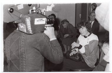 B&W photo of a woman, who is seated. She is facing a television camera operator, who is filming her with the television camera poised on his shoulder. Behind her, there are a a few other people sitting on chairs and looking on. One woman is in the process of sitting on a chair. In the background, there is another woman standing.
