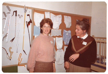 B&W photo of two women standing in front of some display boards affixed to the wall. On the board on the left, various arthritic aids are on display. On the board on the right, a range of underwear and wash cloths are on display.