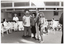 B&W photo of four people, standing, posing for a photo in a large room. Two of the people are in costume, while the other two are dressed in regular clothing. The two people in costume are dressed as the characters Norm and Libby from the government's “Life. Be In It" health promotion campaign. Behind the group, there are some other people sitting on chairs placed in a semi-circle, with one person standing, and an A-frame sandwich board with the heading, 'MIND YOUR BACK'. In the foreground, there is one other person standing to the right of the frame, (partially obscured).