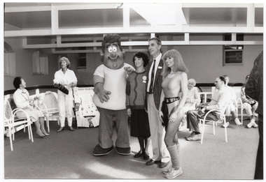 B&W photo of four people, standing, posing for a photo in a large room. Two of the people are in costume, while the other two are dressed in regular clothing. The two people in costume are dressed as the characters Norm and Libby from the government's “Life. Be In It" health promotion campaign. Behind the group, there are some other people sitting on chairs placed in a semi-circle, with one person standing, and an A-frame sandwich board with the heading, 'MIND YOUR BACK'. In the foreground, there is one other person standing to the right of the frame, (partially obscured).