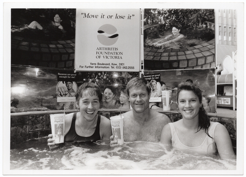 B&W photo of a man and two women sitting in a hot tub/spa. Two of them are holding up PhysiCal branded take-away drink cups. Behind them, there is a display board with a sign and some photos of people in warm water (such as a hot tub/spa or a swimming pool). The sign is titled, "Move it or lose it", followed by a logo and the text "ARTHRITIS FOUNDATION OF VICTORIA Yarra Boulevard, Kew, 3101 For Further Information: Tel. (03) 862.2555". On either side of the sign, there is a photo of a woman in a not tub/spa. A third photo (directly below the sign) depicts the same woman in a spa with two children (one child is obscured). A fourth photo (right) depicts a small group of people partaking in warm water exercises in a swimming pool. There are two small photos superimposed onto the larger photos (each depicting a close up of a man's face) with the text, "Bananas are good enough for me".