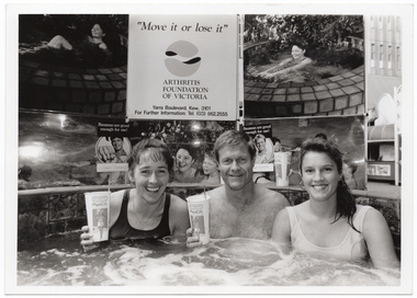 B&W photo of a man and two women sitting in a hot tub/spa. Two of them are holding up PhysiCal branded take-away drink cups. Behind them, there is a display board with a sign and some photos of people in warm water (such as a hot tub/spa or a swimming pool). The sign is titled, "Move it or lose it", followed by a logo and the text "ARTHRITIS FOUNDATION OF VICTORIA Yarra Boulevard, Kew, 3101 For Further Information: Tel. (03) 862.2555". On either side of the sign, there is a photo of a woman in a not tub/spa. A third photo (directly below the sign) depicts the same woman in a spa with two children (one child is obscured). A fourth photo (right) depicts a small group of people partaking in warm water exercises in a swimming pool. There are two small photos superimposed onto the larger photos (each depicting a close up of a man's face) with the text, "Bananas are good enough for me".