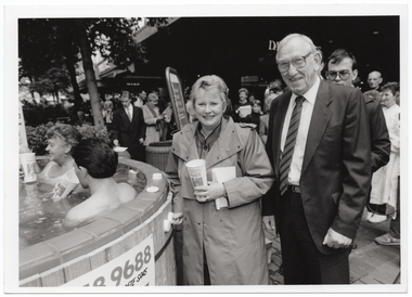 B&W photo of a man and a woman standing beside a hot tub/spa in an outdoor area. There are a few people in the spa, and the woman is pressing the start button on the edge of the spa. She is holding a PhysiCal milkshake cup in her other hand. There is a large crowd of people standing and watching in the background, with the signs of retail department stores, Myer and David Jones, hanging from the plasterboard ceiling above.