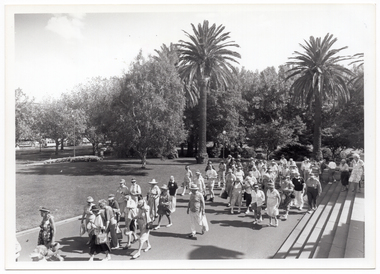 B&W photo of a large group of people walking along a concrete path and down some wide steps in a park. There are two tall palm trees amongst other trees in the park, and there is a tram on the road in the distance (to the left of the frame). The photo has been taken at an angle from above, with the walkers below.