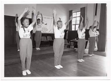 B&W photo of a group of people performing an exercise demonstration in a hall. Hanging up on the wall behind them is a large banner containing the text "Move it or lose it" and "Arthritis Foundation of Victoria", with a logo in between. To one side, there is an information table with boxes underneath and an A-frame sign in front.