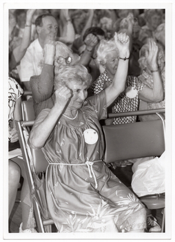 B&W close up photo of a woman sitting on a seat in a row of seats. There is a large crowd of people, also sitting in rows of seats, behind her. Everyone has their arms up, with fists clenched.