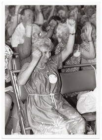 B&W close up photo of a woman sitting on a seat in a row of seats. There is a large crowd of people, also sitting in rows of seats, behind her. Everyone has their arms up, with fists clenched.