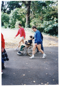 COL photo of a group of people going for a walk along an asphalt path in a park. One of the group is a child who is a wheelchair user. Three adult men are walking alongside him. One man (partially obscured) is pushing the wheelchair, and the other two men are walking on either side of them.