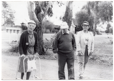 B&W photo of three people walking in or beside a park or reserve. One of them is using a walking frame.