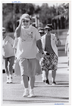 B&W photo of a group of people walking. In the foreground is a woman holding the tip of her sun visor and smiling.