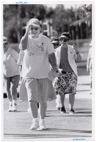 B&W photo of a group of people walking. In the foreground is a woman holding the tip of her sun visor and smiling.