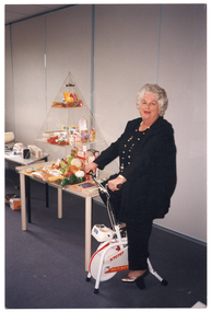 COL photo of a woman sitting on an exercise bike. Beside the exercise bike is a table, on top of which is a clear perspex pyramid-shaped display shelf along the wall. The shelves contain a range of fresh and packaged food products. On another table further along the wall, there are various gadgets or small pieces of equipment on display.
