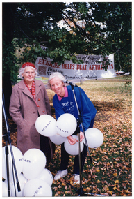 COL photo of two women, standing in a park, behind two microphones stands. One of the women is wearing a 'Move It Or Lose It' windcheater/sweatshirt. She is holding a bunch of white balloons printed with the 'MYER' retail store logo, and there is a large bunch of similar balloons on the ground. In the background, there is a large banner attached to some trees. It contains the text (partially obscured) "...ion of Victoria Ph: 03 862 2555 EXERCISE HELPS BEAT ARTHRITIS Funded by Vi... Promotion Foundation", and the VicHealth logo.