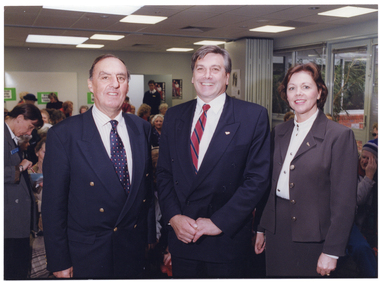 COL photo of three people standing in a row, posing for a photo. Behind them is a room full of people, seated, while a few people are standing. In the background, some posters can be seen on the walls.