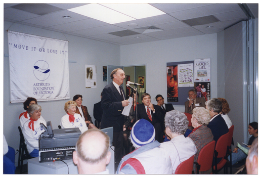 COL photo of a man, standing, addressing an audience. There are several other people seated behind him. On the wall behind them is a large white and blue banner with the text "MOVE IT OR LOSE IT", "ARTHRITIS FOUNDATION OF VICTORIA" and logo. There are various posters on the walls and a display board, including a poster of a dancer.