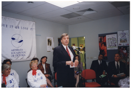 COL photo of a man, standing, addressing an audience. There are several other people seated behind him. On the wall behind them is a large white and blue banner with the text "MOVE IT OR LOSE IT", "ARTHRITIS FOUNDATION OF VICTORIA" and logo. There are various posters on the walls and a display board, including a poster of a dancer.