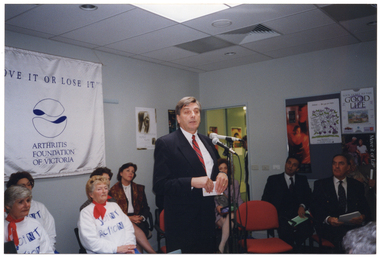 COL photo of a man, standing, addressing an audience. There are several other people seated behind him. On the wall behind them is a large white and blue banner with the text "MOVE IT OR LOSE IT", "ARTHRITIS FOUNDATION OF VICTORIA" and logo. There are various posters on the walls and a display board, including a poster of a dancer.