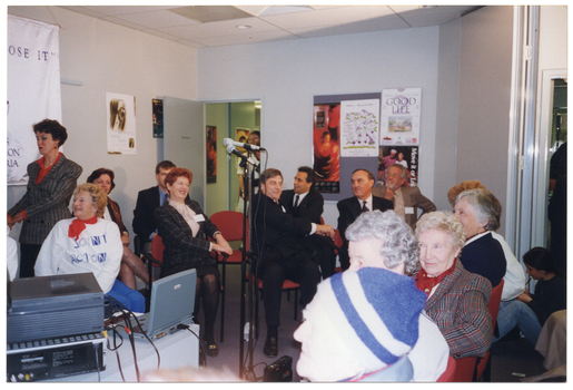 COL photo of a woman, standing. She is holding the hands together in front as she twists her body to one side. On the wall behind her is a large white and blue banner with the text, "MOVE IT OR LOSE IT", "ARTHRITIS FOUNDATION OF VICTORIA", and a logo. In the room, there is a group of people sitting on chairs. They are doing a similar movement with their bodies while remaining seated. A few people are looking in from outside a doorway.  There are various posters on the walls and a display board, including a poster of a dancer.