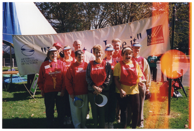 COL photo of a group of people posing for a photo in a park. They are wearing red vests with 'Marshall' printed in white on the front, and two of them are holding a blue and white sunvisor in one hand. The group is standing in front of a large banner. On the banner, there is the text, 'Move It Or Lose It' in large letters across the centre, and logos for the Arthritis Foundation of Victoria and VicHealth Foundation on either side. Behind the banner, there is a large marquee on one side, and a few people (partially obscured) standing behind the edge of the banner at the other end.