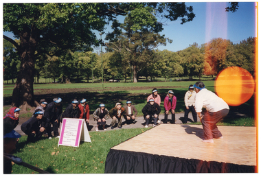 COL photo of a group of people taking part in some Tai Chi exercises in a park. One person on a small stage leads the group in the exercise routine, while the group is crouching down on the grass. Everyone in the group is wearing a blue and white sunvisor, and some people have a head scarf wrapped around the sunvisor. In front of the stage, there is an a-frame sandwich board with various sponsors' names printed on it.
