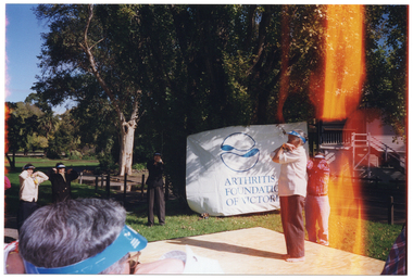 COL photo of a group of people taking part in some Tai Chi exercises in a park. One person on a stage leads the group in the exercise routine, while the people in the group stand on the grass with their hands on their shoulders. Behind them, there is a large blue and white banner with the text "ARTHRITIS FOUNDATION OF VICTORIA" and logo. The banner has been secured to some trees and a wooden building with some rope.