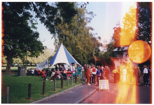 COL photo of a group of people gathered in a park. Some people are sitting on chairs placed around a large blue and white marquee, while others are standing on the grass or on a concrete path and driveway leading to a weatherboard building. There are also a few people gathered inside the marquee, under which there are some rectangular tables. Some people are wearing a red vest with "MARSHALL" in white text. Many are wearing blue and white sunvisors. There is an A-frame sandwich board on the driveway, containing the text, "Ring the Bells for Arthritis - Arthritis Week Joint Walk". To the left of the marquee, there is a large white and blue "Move it or Lose it" banner. Behind the marquee, there is a large white and blue "ARTHRITIS FOUNDATION OF VICTORIA" banner. There is some discolouration on the right hand side of the Photographic image, in the form of a bright orange circle and some vertical lines.