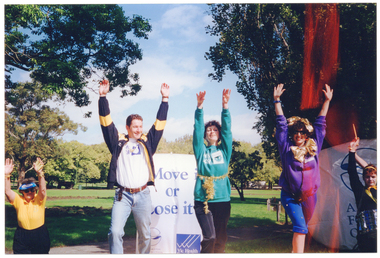COL photo of three people demonstrating some exercises in a park. They are lunging forward while stretching both arms up above their heads. One woman has a strand of gold tinsel tied around her waist, while the other woman has two large gold bows tied around her head and her neck. All three are wearing 'Move It Or Lose It' sweatshirts. Standing on either side of them are two people who are following the exercise routine. One is wearing a blue and white sunvisor while the other is holding a drumstick in her hand and has a small drum hanging from a strap around her neck. Directly behind them is a "Move it or Lose it" freestanding vertical banner, containing the "Arthritis Foundation of Victoria" and "VicHealth Foundation" text and logos. Behind them, to the right of the image, is a large "ARTHRITIS FOUNDATION OF VICTORIA" banner.