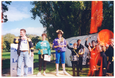 COL photo of three people standing on an outdoor stage in a park. One woman is holding a large piece of paper or booklet, while the other woman is holding a small piece of paper. All three are wearing 'Move It Or Lose It' sweatshirts. To their left are members of a drum marching band. Some of the drummers are holding one drumstick up in the air. Directly behind them is a "Move it or Lose it" freestanding vertical banner, containing the Arthritis Foundation of Victoria" and VicHealth Foundation text and logos. Behind the marching band is a large "ARTHRITIS FOUNDATION OF VICTORIA" banner.