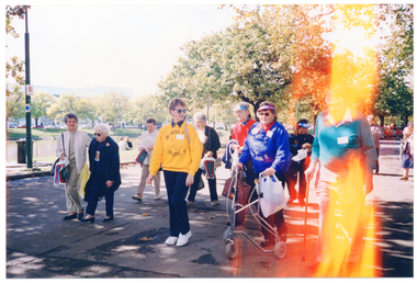 COL photo of a group of people walking along a concrete or asphalt road lined with trees. In the background, there is a river, with some buildings on the other side of the bank. Two women in the foreground are wearing 'MOVE IT OR LOSE IT' sweatshirts. One of them is using a walking frame. A man walking next to her is using a walking stick. Some people are wearing blue and white sunvisors, and several people are carrying the same type of promotional plastic bag.