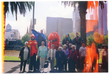 COL photo of a group of people posing for a photo on the riverbank with some stiltwalkers in colourful costumes. Behind them is a river with some buildings visible on the bank on other side of the river, including some high rise buildings and a church. A digital clock displays the time as being "12.32". It is flanked by advertisements for Samsung Electronics and Crown Casino.