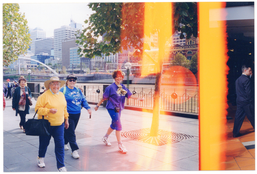 COL photo of a group of people walking along a path by a river bank. In the background, there are some large and high-rise buildings on the other side of the river and a bridge connecting the two riverbanks.