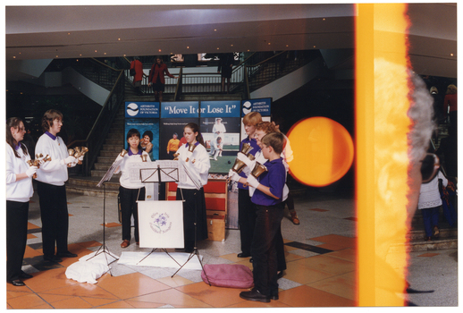COL photo of a troupe of performers holding various sized handbells. They are standing in a semi-circle formation in an outdoor forecourt of a building. Behind them is a large freestanding multipanel display board containing the text "Move It or Lose It" in the centre, and "Arthritis Foundation of Victoria" on either side. In the background, there is a set of stairs leading up to the first floor of the building, with several people walking up and down the staircase.