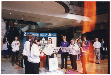 COL photo of a troupe of performers holding various sized handbells. They are standing in a semi-circle formation in an outdoor forecourt of a building. Behind them is a large freestanding multipanel display board containing the text "Move It or Lose It" in the centre, and "Arthritis Foundation of Victoria" on either side. In the background, there is a set of stairs leading up to the first floor of the building, with several people walking up and down the staircase. A woman standing in front of the staircase is wearing a 'Move It or Lose It" sweatshirt.  There are several people walking around the ground floor, behind the performers.