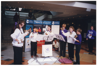 COL photo of a troupe of performers holding various sized handbells. They are standing in a semi-circle formation in an outdoor forecourt of a building. Behind them is a large freestanding multipanel display board containing the text "Move It or Lose It" in the centre, and "Arthritis Foundation of Victoria" on either side. In the background, there is a set of stairs leading up to the first floor of the building, with several people walking up and down the staircase. There are several people walking around the ground floor, and some watching the performers.