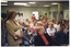 COL photo of a room full of people sitting on rows of chairs. One woman is standing in the foreground, to the left of the frame, and a few other people are standing at the back of the room. Behind the woman in the foreground, there is a camera operaor behind a television camera on a stand (partially obscured). On the walls around the room, there are several posters depicting the image of the woman standing in the foreground.
